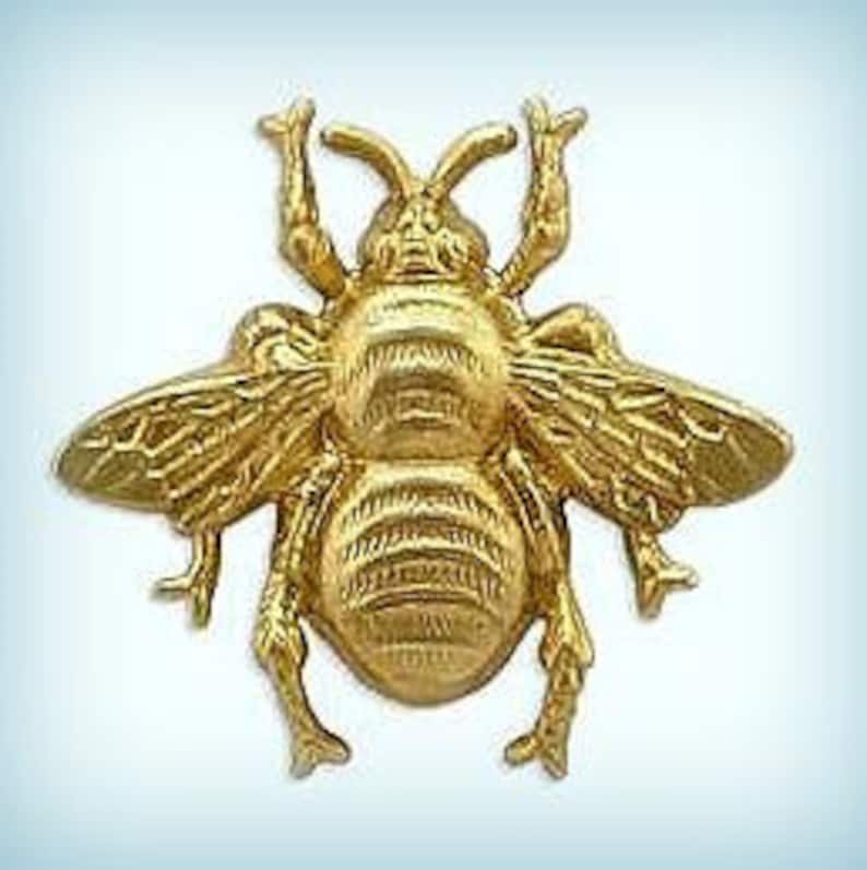 May include: A gold-coloured metal bee figurine with detailed wings and legs. The bee is in a flying position with its wings spread out.