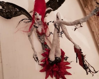 May include: A handmade fairy doll with red hair, black wings, and a red and black skirt. The doll is posed with its legs raised and its arms outstretched. The doll is made of fabric and other materials. The doll is signed "Botanik Art Studio©".