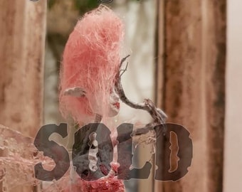 May include: A pink and white fairy doll with a pink cotton candy-like hairdo and a branch in her hand. The doll is wearing a pink and white dress and has a white face with black eyes. The doll is standing in front of a mirror with the word "SOLD" written in black letters over the image.