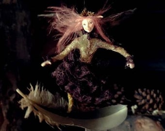 May include: A whimsical fairy figurine with pink hair and a brown dress, perched on a crescent moon with a feather attached. The fairy is holding a small branch in its hand. The background is dark and blurry, with the text "Botanic Tales©" at the top.