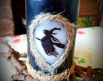 May include: Black pillar candle with a silhouette of a witch flying on a broomstick inside a burlap heart. The candle is on a wooden base with the text "Botanic Tales©".