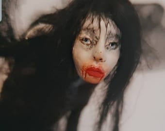 May include: A doll with long black hair and a white face. The doll has red lipstick and a small amount of red paint on its chin, resembling blood.