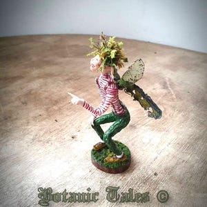 May include: A whimsical fairy figurine with green moss hair, a red and white striped shirt, green trousers, and paper wings. The fairy is sitting on a small green base.