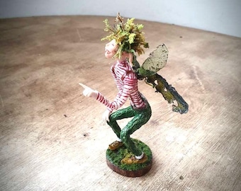 May include: A whimsical fairy figurine with green moss hair, a red and white striped shirt, green pants, and paper wings. The fairy is sitting on a small green base.