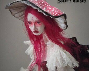 May include: A porcelain doll with long pink hair wearing a white dress with lace trim and a large red and white mushroom hat. The doll's face is painted with delicate features and rosy cheeks. The hat has a white underside and a red top with white polka dots. The doll is standing on a red and white polka dot base.