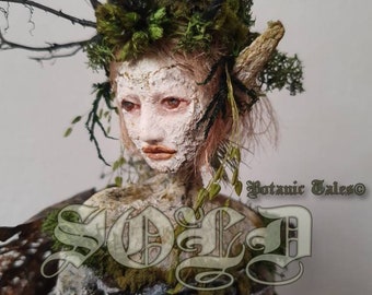 May include: A handmade doll with a mossy green crown, a white face, and a birch bark body. The doll is wearing a green moss dress and has a twig-like horn on its head. The doll is titled "Botanic Tales".