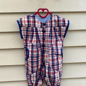 Può includere: Un pagliaccetto vintage a quadri rossi, bianchi e blu, con finiture e tasche blu. Il pagliaccetto ha una fodera a quadretti blu e bianca allo scollo e un piccolo disegno ricamato. È appeso a un gancio a forma di cuore rosa.