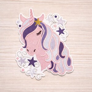 Könnte beinhalten: Gestickter Aufnäher mit einem rosa Einhornkopf mit goldenem Horn und Stern. Das Einhorn hat lila und silbernes Haar, geschlossene Augen und ist von Sternen und Wirbeln umgeben. Der Aufnäher befindet sich auf weißem Hintergrund.