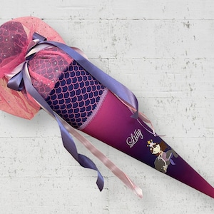 Könnte beinhalten: Ein rosa-lilafarbener Schultüte mit einem Meerjungfrauen-Motiv und dem Namen "Lilly" darauf. Die Tüte ist mit einer rosa Tüllschleife und einem lila Band verziert.