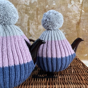 以下が含まれることがあります： グレーのポンポンが付いた、2つのニットティーコージー。 コージーは、グレー、ラベンダー、ネイビーブルーのセクションでリブ編みになっています。 ティーコージーは茶色のティーポットの上に置かれています。