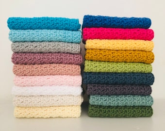 May include: A stack of 10 knitted dishcloths in various colours, including blue, pink, yellow, green, and grey. The dishcloths are stacked on a white surface.