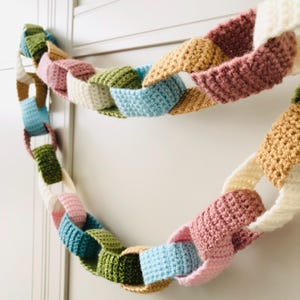 May include: A decorative garland made of interlocking crocheted fabric rings in various colors including cream, dusty pink, mustard yellow, light blue, olive green, and teal. The chain consists of multiple soft, textured loops linked together.