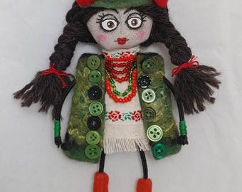 Puede incluir: Muñeca de tela hecha a mano con vestimenta tradicional. La muñeca presenta un chaleco verde adornado con botones, una blusa blanca bordada y un collar de cuentas rojas. Tiene cabello oscuro, detalles rojos y botas rojas. La muñeca mide unos 15 cm de altura.