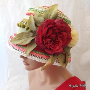 May include: A multi-coloured straw bucket hat featuring horizontal stripes in shades of pink, green, yellow, and white. The hat is decorated with a large dark red fabric peony, green fabric leaves, and a row of small olive green pom-poms.