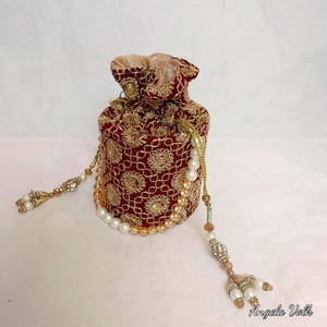 May include: A burgundy drawstring pouch with gold floral embroidery. The bag features a gold drawstring and is adorned with pearl and crystal beaded tassels. The interior lining is a light gold colour.