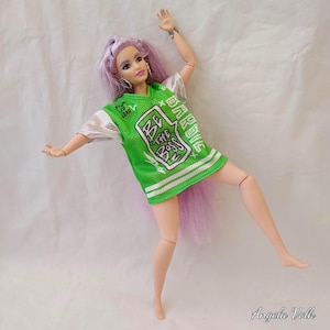 May include: A doll with long lavender hair wearing a bright green mesh jersey with silver metallic sleeves. The jersey features white text that reads "BE THE BOSS" and "BORN TO LEAD" with "BARBIE" printed vertically on the side in white.