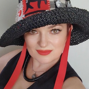 Può includere: Un cappello a secchiello nero intrecciato con una toppa in tessuto rosso raffigurante un gatto nero, inserti in tessuto scozzese bianco e nero, bottoni rossi a forma di cuore e lunghi nastri rossi. Il testo newly decorated hat è visibile.