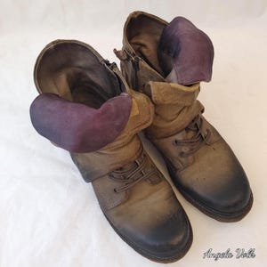 May include: A pair of mid-calf leather boots featuring a distressed olive-brown finish with dark charcoal toe caps. The boots include lace-up fronts, side zippers, and distinctive folded cuffs lined with a deep purple fabric interior.