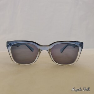May include: Blue-tinted sunglasses with a square frame. The lenses are dark grey, and the frame is a translucent blue. The sunglasses are on a light beige surface. The name "Angela Volk" is in the bottom right corner.