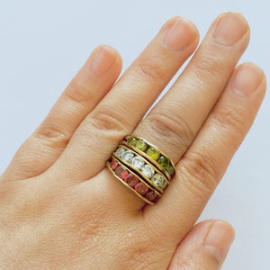 Può includere: Un anello dorato con quattro file di pietre preziose colorate. Le file presentano pietre rosse, bianche e verdi. L'anello è indossato su un dito, mettendo in risalto il design e i colori del gioiello.