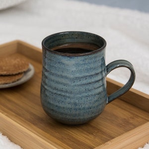 Könnte beinhalten: Eine blaue Keramiktasse gefüllt mit Kaffee steht auf einem Holztablett mit zwei runden Keksen. Das Tablett befindet sich auf einer weißen, flauschigen Oberfläche.