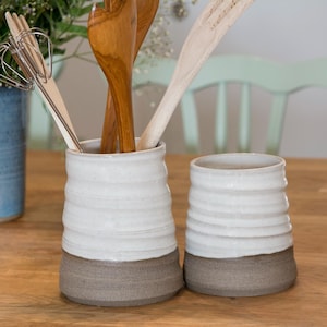 Puede incluir: Dos portautensilios de cerámica blanca con bases marrones. Los portautensilios están llenos de cucharas de madera y un batidor.