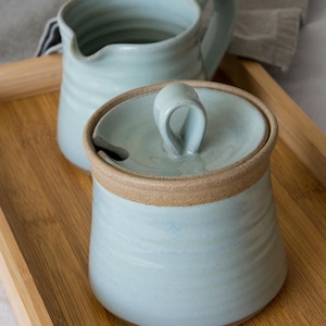 May include: A light blue ceramic sugar bowl with a matching lid featuring a loop handle and a small notch for a spoon, accompanied by a matching creamer jug. Both pieces have a ribbed texture and an exposed brown clay rim at the top.