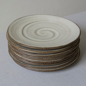 Puede incluir: Una pila de platos de cer&aacute;mica blanca con un borde marr&oacute;n y un dise&ntilde;o en espiral. Los platos est&aacute;n apilados sobre un mantel blanco.