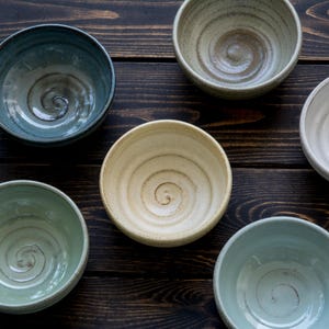 Op de afbeelding: Zes keramische kommen in verschillende kleuren: blauw, groen en crème. Elke kom heeft een spiraalvormig patroon in het midden. De kommen zijn gerangschikt op een donkerbruin houten oppervlak.
