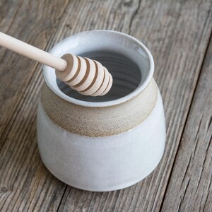 Puede incluir: Un tarro de miel de cerámica con cuerpo esmaltado en blanco y borde de color tostado natural, con una abertura ancha. Un cazo para miel de madera clara con cabeza ranurada está colocado en diagonal, extendiéndose hacia el centro del recipiente.