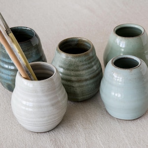 Puede incluir: Conjunto de cinco jarrones de cerámica en tonos azules, verdes y blancos. Un jarrón contiene pinceles. Los jarrones tienen forma redondeada con superficie texturizada y acanalada. Ideal para decoración del hogar o como regalo único.