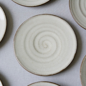 Puede incluir: Juego de seis platos de cer&aacute;mica blanca con un dise&ntilde;o en espiral. Los platos tienen un borde marr&oacute;n y est&aacute;n dispuestos en un patr&oacute;n circular sobre una superficie gris claro.