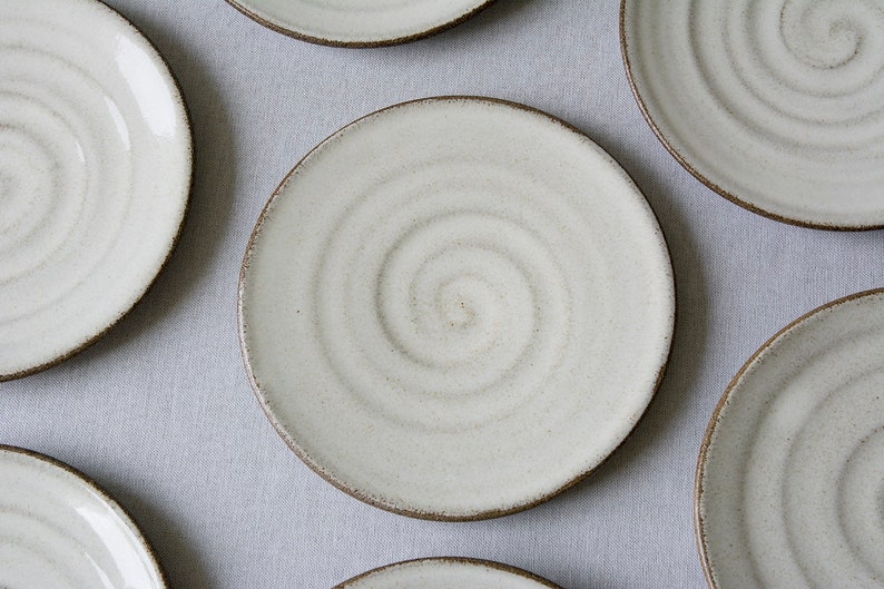 Puede incluir: Juego de seis platos de cer&aacute;mica blanca con un dise&ntilde;o en espiral. Los platos tienen un borde marr&oacute;n y est&aacute;n dispuestos en un patr&oacute;n circular sobre una superficie gris claro.