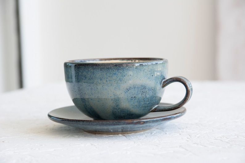 May include: A blue ceramic teacup and saucer set. The cup has a speckled glaze and a rounded shape. The saucer is a simple, round shape.