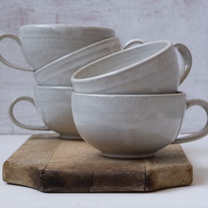 May include: A stack of four white ceramic mugs with rounded handles on a wooden cutting board. The mugs are stacked in a staggered pattern, with the top mug slightly offset from the others.