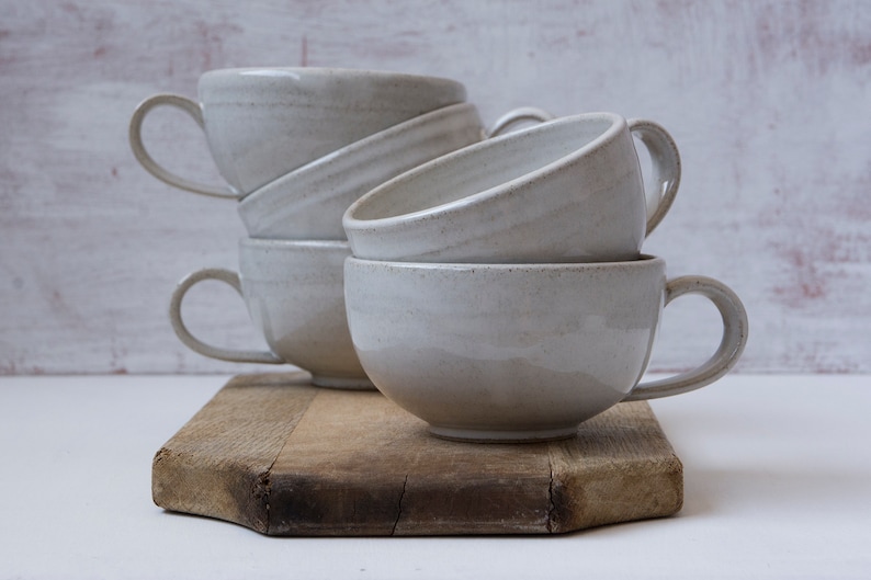 May include: A stack of four white ceramic mugs with rounded handles on a wooden cutting board. The mugs are stacked in a staggered pattern, with the top mug slightly offset from the others.