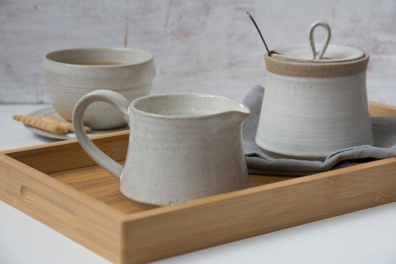 Op de afbeelding: Een witte keramische kan, suikerpot en theekopje op een houten dienblad. De kan heeft een handvat en een tuit. De suikerpot heeft een deksel met een knop. Het theekopje is gevuld met thee.