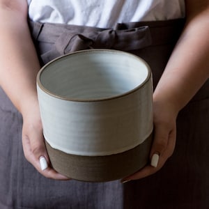 以下が含まれることがあります： 手作りの陶器プランター。上部はクリームホワイト、下部はウォームブラウンのツートンカラー。円筒形で、ミニマルなデザイン。人が持っており、その形状とデザインが際立っています。