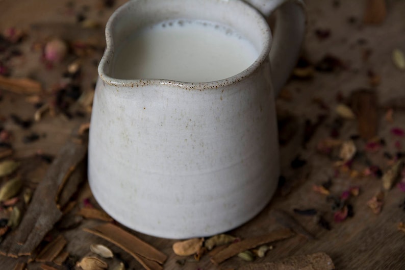 Op de afbeelding: Een witte keramische kan gevuld met melk, die op een oppervlak staat dat bedekt is met bruine specerijen.