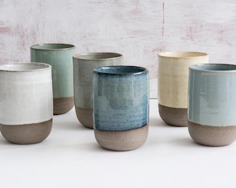 May include: Six ceramic mugs in various shades of blue, green, and white with a brown rim. The mugs are arranged in a row on a white surface.