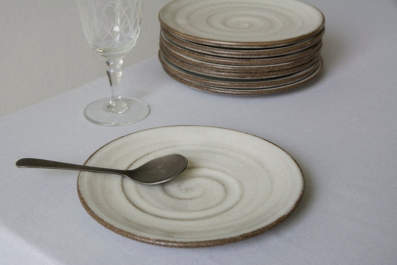 Puede incluir: Un plato de cer&aacute;mica blanco con un dise&ntilde;o en espiral y un borde marr&oacute;n. Una cuchara de plata descansa sobre el plato. El plato est&aacute; sobre un mantel blanco.