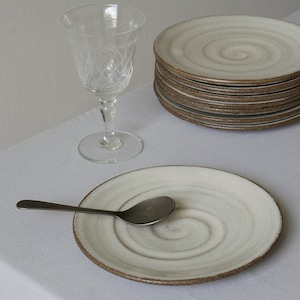 Puede incluir: Un juego de platos de cer&aacute;mica blanca con un dise&ntilde;o en espiral y un borde marr&oacute;n. Los platos est&aacute;n apilados uno encima del otro. Una cuchara de plata est&aacute; en un plato. Un vaso transparente est&aacute; en el fondo.