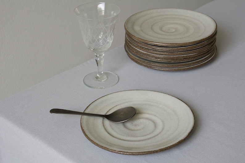 Puede incluir: Un juego de platos de cer&aacute;mica blanca con un dise&ntilde;o en espiral y un borde marr&oacute;n. Los platos est&aacute;n apilados uno encima del otro. Una cuchara de plata est&aacute; en un plato. Un vaso transparente est&aacute; en el fondo.