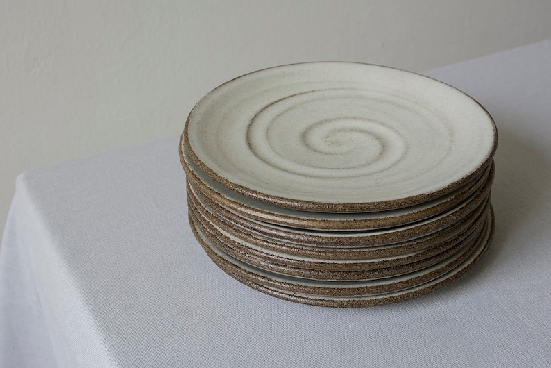 Puede incluir: Una pila de platos de cer&aacute;mica blanca con un borde marr&oacute;n y un dise&ntilde;o en espiral. Los platos est&aacute;n apilados sobre un mantel blanco.
