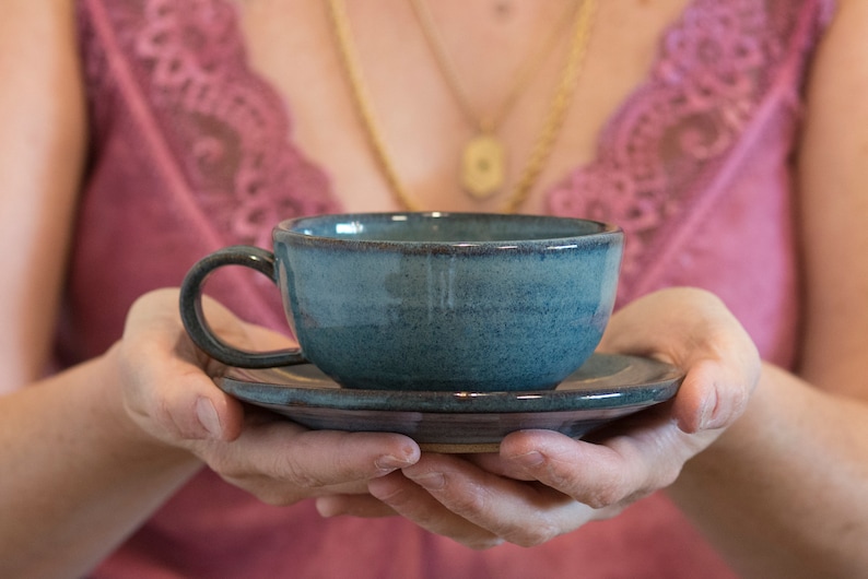 May include: A person is holding a blue ceramic teacup and saucer. The cup and saucer have a speckled glaze.