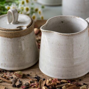 Op de afbeelding: Een witte keramische roomkan en suikerpot set met een bruine rand. De roomkan heeft een handvat en de suikerpot heeft een deksel. De set staat op een houten oppervlak met gedroogde kruiden en specerijen.