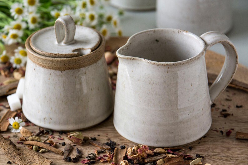 Op de afbeelding: Een witte keramische roomkan en suikerpot set met een bruine rand. De roomkan heeft een handvat en de suikerpot heeft een deksel. De set staat op een houten oppervlak met gedroogde kruiden en specerijen.