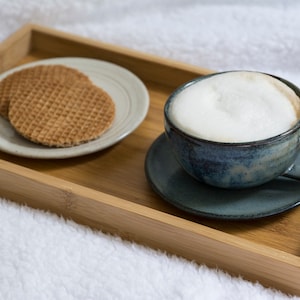 May include: A wooden tray with a cup of coffee and two waffle cookies on a white plate. The coffee cup is blue and white with a white foam top. The tray is on a white fluffy surface.