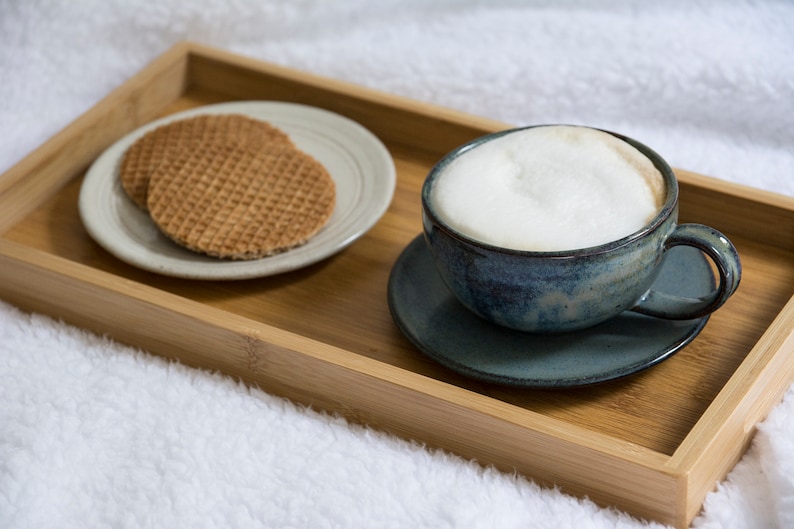 May include: A wooden tray with a cup of coffee and two waffle cookies on a white plate. The coffee cup is blue and white with a white foam top. The tray is on a white fluffy surface.