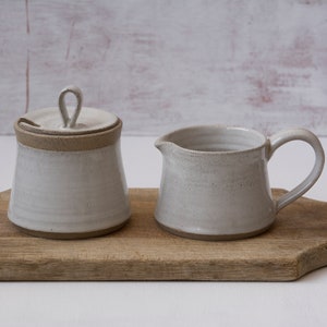 Op de afbeelding: Een witte keramische roomkan en suikerpot set met een deksel op een houten snijplank. De roomkan heeft een handvat en de suikerpot heeft een deksel met een klein handvat.